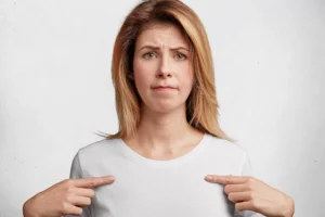 A woman with shoulder-length blonde hair wears a plain white T-shirt and points to her chest with both hands. She has a slightly furrowed brow and a concerned, uncertain expression, suggesting discomfort or dissatisfaction—possibly related to how her bra fits or feels.