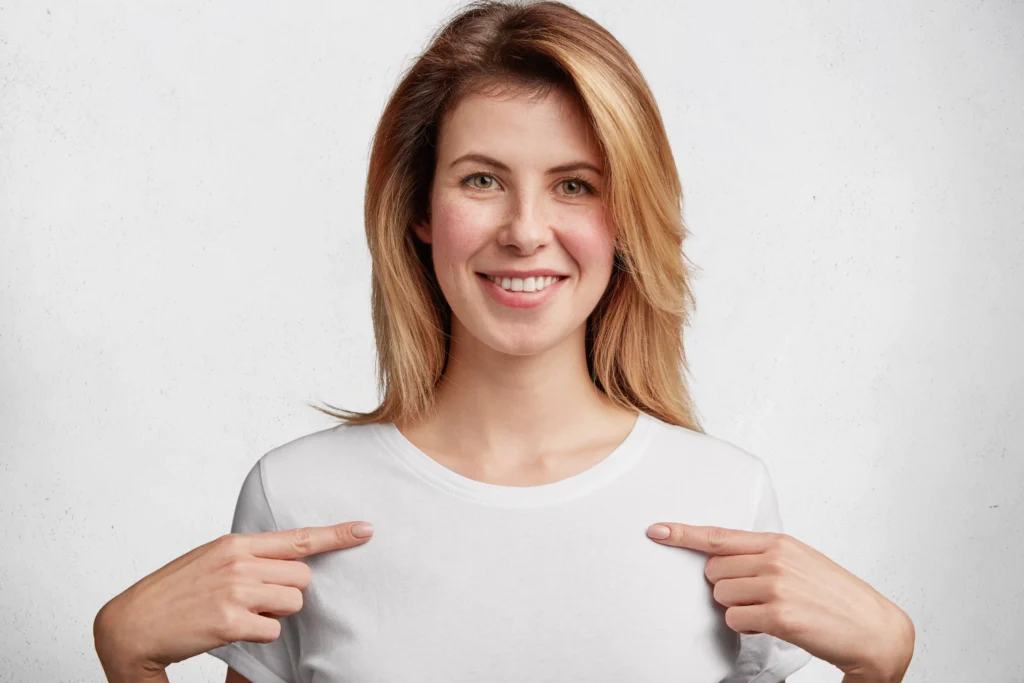 The same woman, now smiling confidently, points to her chest with both hands. Her expression is happy and self-assured, suggesting she feels supported and comfortable—likely due to wearing a properly fitting bra.