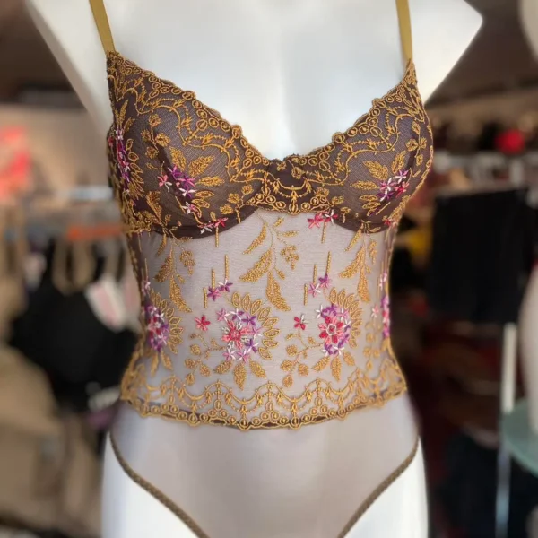 A mannequin wearing a sheer bodysuit in a warm brown color with intricate floral embroidery in gold and pink. The bodysuit has delicate lace detailing along the cups and torso, with thin shoulder straps. The embroidered floral patterns add an elegant, decorative touch to the design.