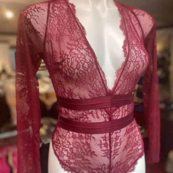 A burgundy lace long-sleeve bodysuit displayed on a mannequin. The bodysuit features a plunging V-neckline, sheer floral lace fabric, and two solid horizontal bands around the waist, adding structure to the delicate lace design.